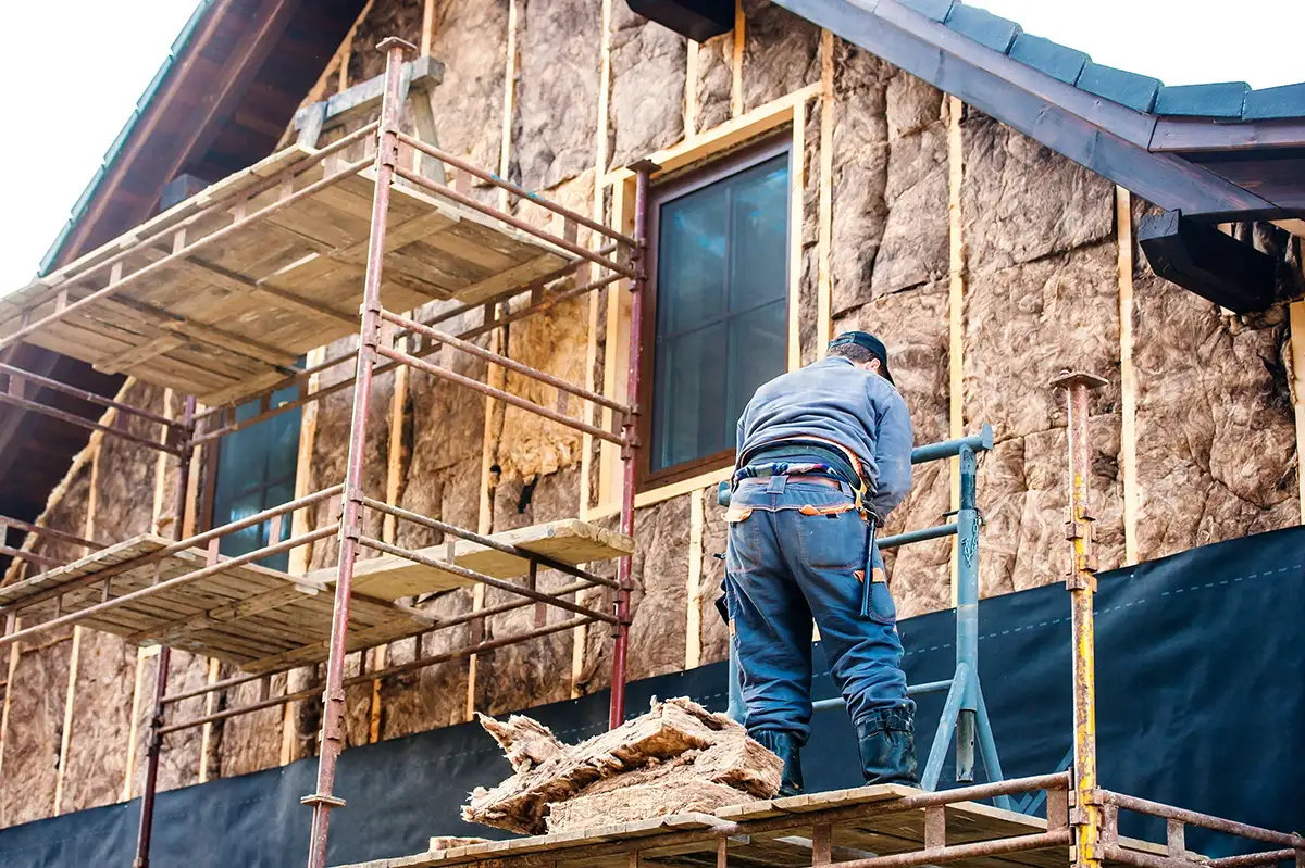 En byggearbejder står på stillads og installerer isolering på ydervæggen af et hus under renovering.