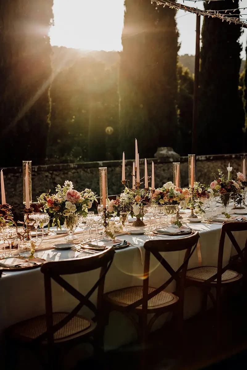 Et udendørs spisebord dækket med blomsterarrangementer, høje lys, tallerkener og glas, omgivet af træstole ved solnedgang.